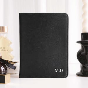 May include: Black leather passport holder with the initials "M.D." in white. The cover has a textured appearance. The passport holder is rectangular and appears to be closed. A candle and perfume bottle are in the background.