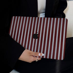 Puede incluir: Una funda para portátil con un patrón de rayas verticales en burdeos, azul claro y blanco. La funda presenta un logotipo de Apple negro y la sostiene una persona que lleva una blazer negra y pantalones negros.