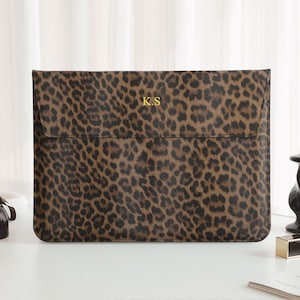 May include: A brown and black leopard print laptop sleeve with the gold initials "K.S." on the front. The sleeve is rectangular with a flap closure. The background includes a white candle, perfume bottles, and a black candle holder.