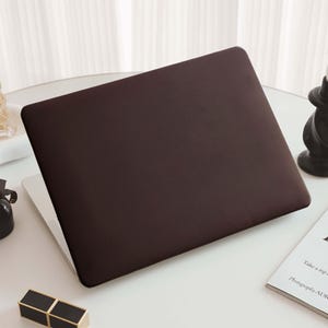 May include: A closed laptop featuring a solid dark brown matte protective cover. The laptop is positioned on a white surface alongside a clear glass perfume bottle, a small black bottle with a ribbon, a black candle holder, and a white book.