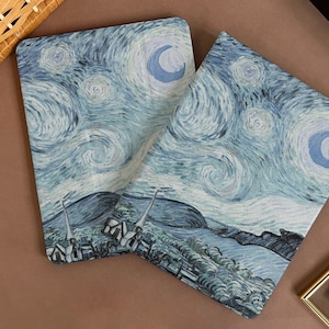 Puede incluir: Dos fundas de cojín azul y blanco con un diseño de remolino que recuerda a un cielo estrellado. Las fundas presentan una luna creciente y un paisaje con árboles y colinas.