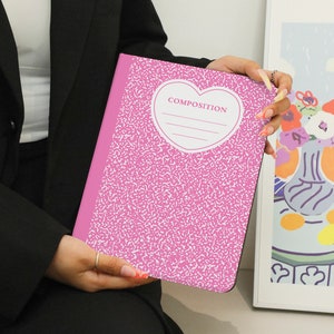 Puede incluir: Cuaderno de composición rosa con un recorte en forma de corazón y la palabra "COMPOSITION" impresa en su interior. El cuaderno tiene un patrón moteado rosa y blanco.