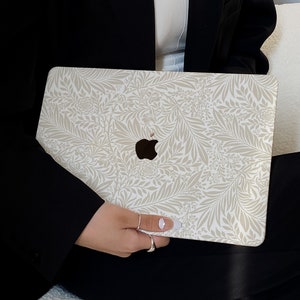Op de afbeelding: Een witte laptop met een beige bloemenpatroon op de deksel. De laptop wordt vastgehouden door een persoon die een zwarte blazer draagt.
