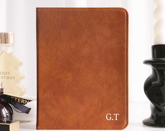 May include: A brown leather journal with the initials "G.T." in white. The journal is rectangular and has a textured surface. The background includes a candle and a perfume bottle.