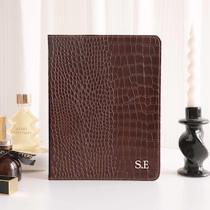 May include: A brown leather tablet case with a crocodile skin texture. The case has the initials "S.E" in white on the lower right corner. A decorative candle and perfume bottles are in the background.