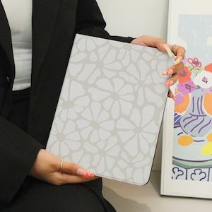 May include: A light gray tablet case featuring a repeating floral pattern in a lighter shade of gray. The case is held by a person wearing a black blazer. The background includes a framed artwork with a floral still life.