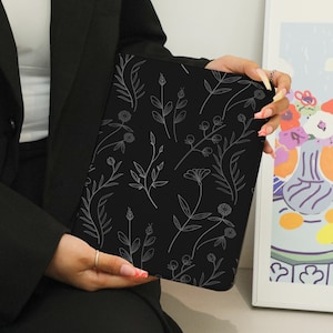 May include: A black laptop sleeve with a white floral pattern. The sleeve is held by a person's hand.