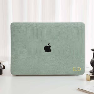 May include: A light green laptop case with a crocodile skin texture. The case has a black Apple logo and gold initials "E.D." on the lower right. The case is on a white surface with decorative items in the background.