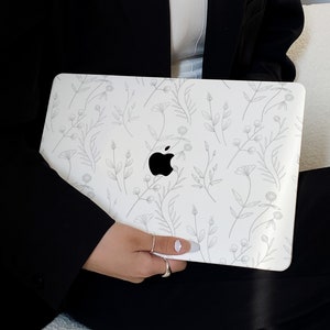 Op de afbeelding: Een witte laptop met een bloemenpatroon en een zilveren Apple-logo. De laptop wordt vastgehouden door een persoon die een zwarte blazer draagt.