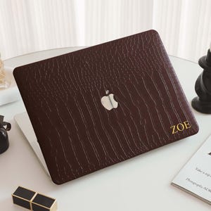 May include: A brown laptop case with a textured pattern and the Apple logo. The name "ZOE" is printed in gold on the lower right corner. The case is on a white surface with a candle and perfume bottles.
