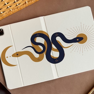 Pode incluir: Uma capa de telefone branca com uma lua crescente dourada e estrelas no lado esquerdo e uma cobra azul com um sol dourado no lado direito.