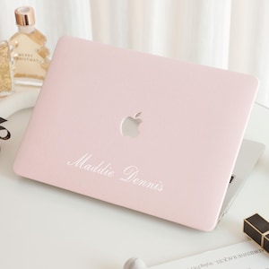 Puede incluir: Un portátil rosa claro con el logotipo de Apple y el nombre "Maddie Dennis" en escritura blanca. El portátil está abierto sobre una superficie blanca, con frascos de perfume y un lápiz labial cerca.