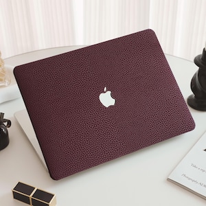 May include: A burgundy laptop with a textured cover and the Apple logo. The laptop is on a white surface, next to a candle, perfume bottles, and a magazine with the text "In the of the".