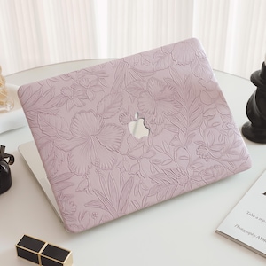 Puede incluir: Una funda para portátil lila claro con un diseño floral en relieve. La funda está abierta, revelando el logotipo plateado de Apple. El diseño presenta flores y hojas grandes. Otros elementos en la mesa incluyen frascos de perfume, una vela y una revista.