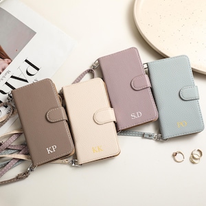 May include: Four leather phone cases in shades of taupe, cream, lavender, and light blue, each with a strap and snap closure. The cases are personalized with initials. A magazine with the word "PARIS" is in the background.