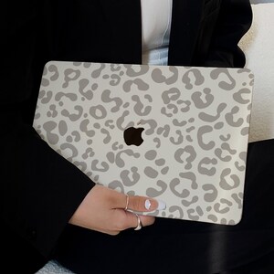 May include: A laptop case featuring a light gray leopard print design. The case has a white background and a cut-out for the Apple logo. The case is held by a person wearing a black blazer and black pants.