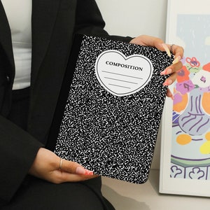 Puede incluir: Una libreta de composición negra y blanca con un recorte en forma de corazón que dice "COMPOSITION".