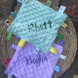 May include: Two square baby security blankets with a dimpled minky texture. The top blanket is light green with the name Rhett embroidered in dark green. The bottom blanket is light purple with the name Baylor embroidered in dark purple. Both feature various colourful patterned ribbon tags along the edges.