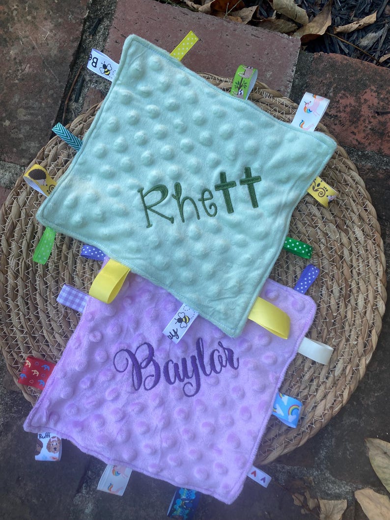 May include: Two square baby security blankets with a dimpled minky texture. The top blanket is light green with the name Rhett embroidered in dark green. The bottom blanket is light purple with the name Baylor embroidered in dark purple. Both feature various colourful patterned ribbon tags along the edges.