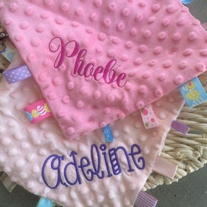 May include: Two square baby security blankets with a dimpled minky texture. The top blanket is bright pink with the name Phoebe embroidered in dark pink. The bottom blanket is light pink with the name Adeline embroidered in purple. Both feature colourful ribbon tags.