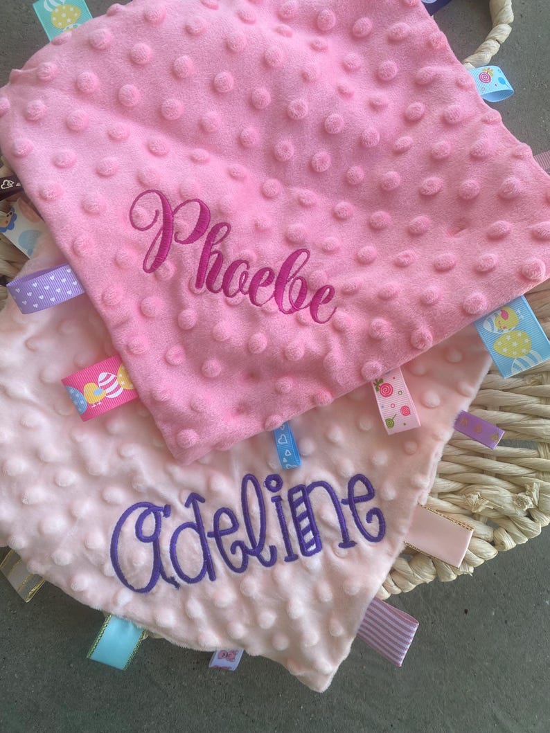 May include: Two square baby security blankets with a dimpled minky texture. The top blanket is bright pink with the name Phoebe embroidered in dark pink. The bottom blanket is light pink with the name Adeline embroidered in purple. Both feature colourful ribbon tags.