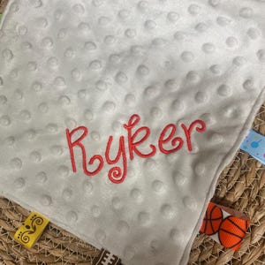 May include: A square white minky fabric security blanket with a dimpled texture. The name Ryker is embroidered in red script in the centre. The edges feature various colourful ribbon tags, including patterns of basketballs, hearts, and geometric shapes.