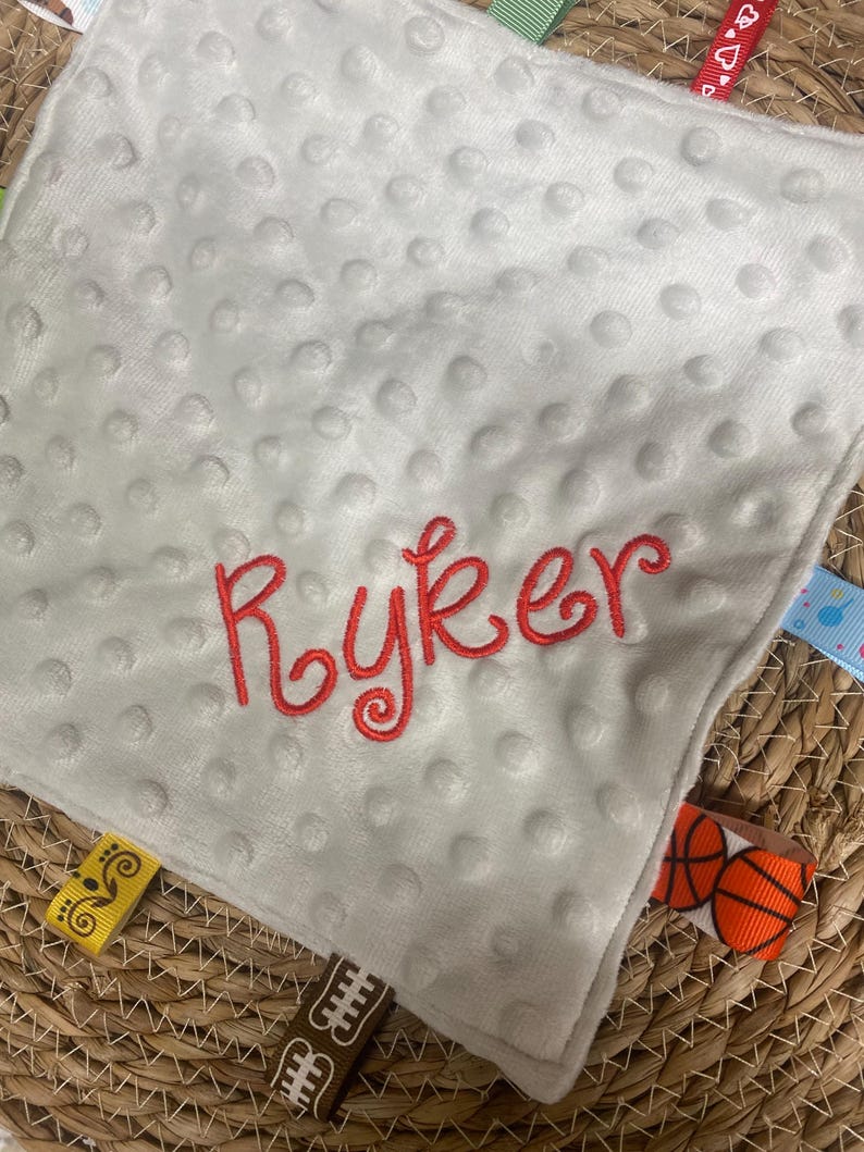 May include: A square white minky fabric security blanket with a dimpled texture. The name Ryker is embroidered in red script in the centre. The edges feature various colourful ribbon tags, including patterns of basketballs, hearts, and geometric shapes.