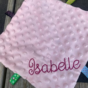 May include: A square, light pink minky fabric security blanket with a raised dimpled texture. The name Isabelle is embroidered in dark pink script across the bottom. Multiple colourful ribbon tags with various patterns extend from the edges.
