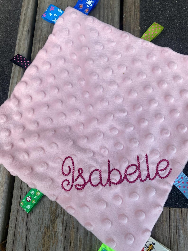 May include: A square, light pink minky fabric security blanket with a raised dimpled texture. The name Isabelle is embroidered in dark pink script across the bottom. Multiple colourful ribbon tags with various patterns extend from the edges.