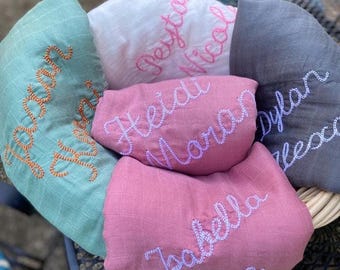 May include: A collection of personalized baby blankets in various colors, including teal, pink, white, and gray. Each blanket features a name embroidered in a contrasting color, such as orange, pink, and blue. The blankets are arranged in a circular pattern.