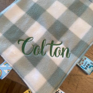 May include: A square baby security blanket featuring a sage green and white buffalo check pattern. The name Colton is embroidered in dark green script in the centre. The blanket has various colourful ribbon tags attached to the edges.