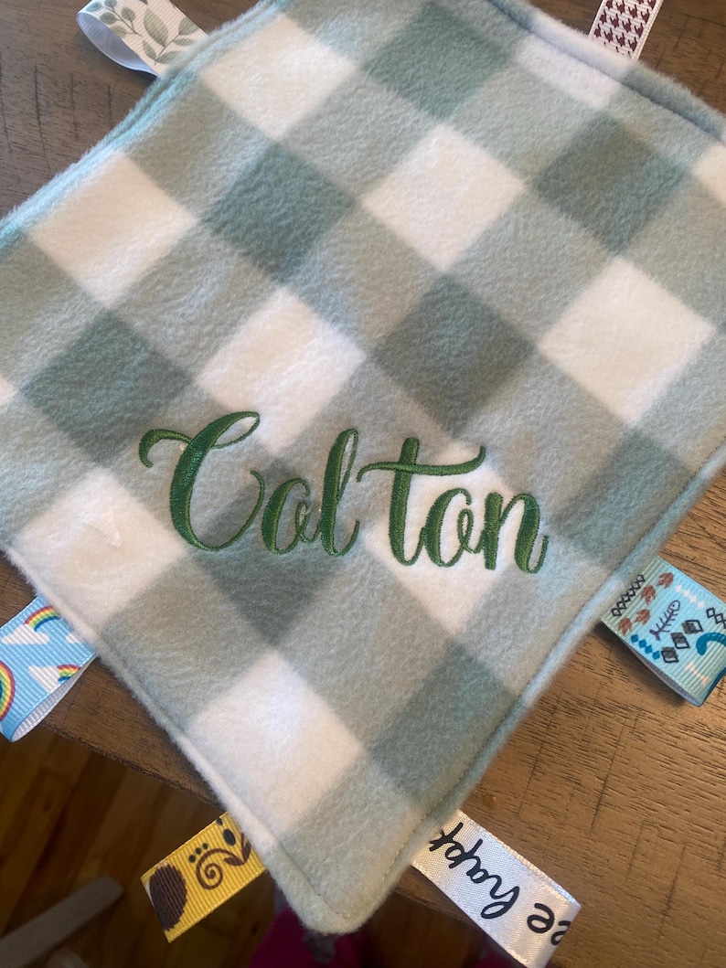 May include: A square baby security blanket featuring a sage green and white buffalo check pattern. The name Colton is embroidered in dark green script in the centre. The blanket has various colourful ribbon tags attached to the edges.
