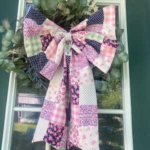 Peut inclure: Une couronne décorative avec un grand nœud patchwork. Le nœud présente une variété de tissus, notamment du vichy rose, des imprimés floraux et des pois bleu marine. La couronne est ornée de feuillage vert et de fleurs blanches, créant un look charmant et artisanal.