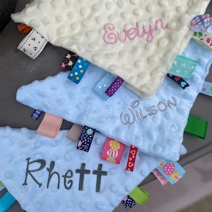 May include: Three triangular baby security blankets with dimpled minky fabric. The top blanket is cream with pink embroidery reading Evelyn. The middle is light blue with grey embroidery reading Wilson. The bottom is light blue with grey embroidery reading Rhett. Each features various colourful patterned ribbon tags.