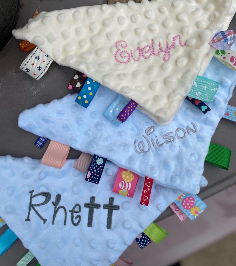 May include: Three triangular baby security blankets with dimpled minky fabric. The top blanket is cream with pink embroidery reading Evelyn. The middle is light blue with grey embroidery reading Wilson. The bottom is light blue with grey embroidery reading Rhett. Each features various colourful patterned ribbon tags.