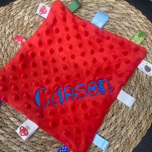 May include: A square red minky fabric security blanket with a dimpled texture. The name Carson is embroidered in blue thread across the centre. The blanket features multiple colourful ribbon tags around the edges, including patterns of mushrooms and dots.