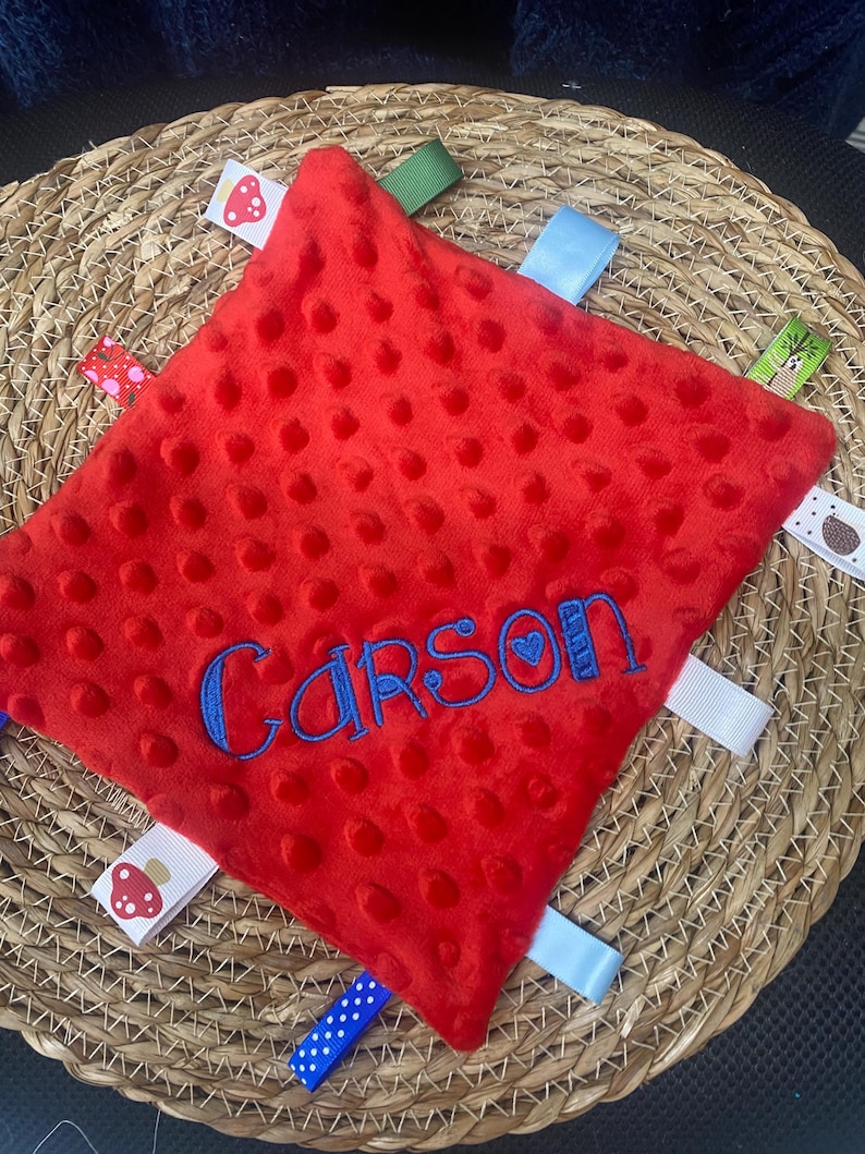 May include: A square red minky fabric security blanket with a dimpled texture. The name Carson is embroidered in blue thread across the centre. The blanket features multiple colourful ribbon tags around the edges, including patterns of mushrooms and dots.