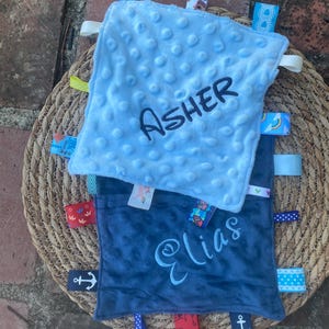 May include: Two square baby security blankets with dimpled minky fabric. The top blanket is light blue with the name Asher embroidered in dark thread. The bottom blanket is dark blue with the name Elias embroidered in light thread. Both feature colourful ribbon tags.