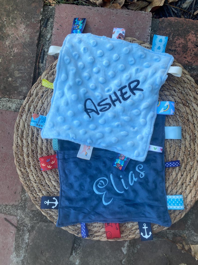 May include: Two square baby security blankets with dimpled minky fabric. The top blanket is light blue with the name Asher embroidered in dark thread. The bottom blanket is dark blue with the name Elias embroidered in light thread. Both feature colourful ribbon tags.