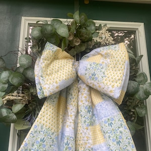 May include: A decorative wreath featuring green eucalyptus leaves and white floral accents, adorned with a large, multi-patterned fabric bow. The bow displays a patchwork design of yellow gingham, small yellow flowers, and blue floral botanical prints.