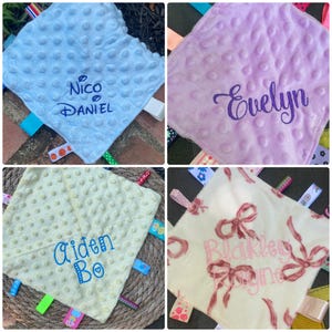May include: A collection of four square sensory blankets featuring dimpled minky fabric in light blue, lavender, and pale yellow, plus one cream blanket with a pink bow print. Each blanket is personalised with embroidered names and colourful ribbon tags.