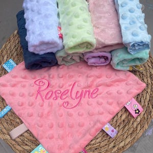 May include: A pink minky fabric security blanket with a dimpled texture, embroidered with the name Roselyne in cursive script. The blanket features multiple colourful ribbon tags along the edges, with a stack of similar blankets in various colours behind it.