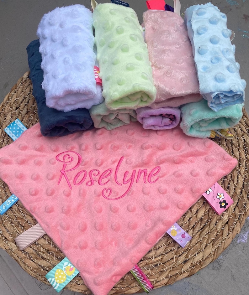 May include: A pink minky fabric security blanket with a dimpled texture, embroidered with the name Roselyne in cursive script. The blanket features multiple colourful ribbon tags along the edges, with a stack of similar blankets in various colours behind it.