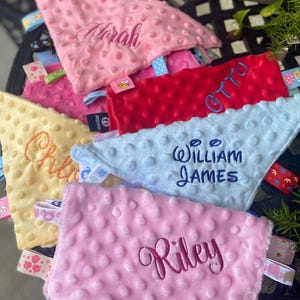 May include: A collection of soft, dimpled minky fabric security blankets in pink, red, light blue, and yellow. Each blanket features embroidered names including Norah, Otto, William James, and Riley, along with various colourful patterned ribbon tags.