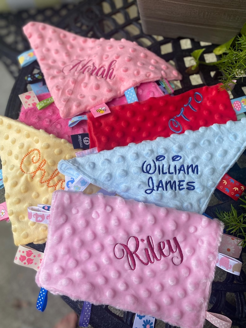 May include: A collection of soft, dimpled minky fabric security blankets in pink, red, light blue, and yellow. Each blanket features embroidered names including Norah, Otto, William James, and Riley, along with various colourful patterned ribbon tags.