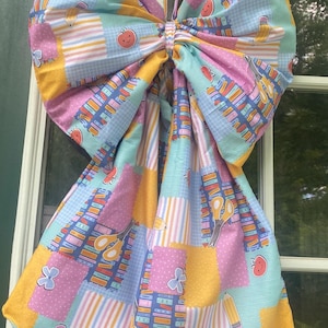 Könnte beinhalten: Eine große Stoffschleife mit einem schulbezogenen Muster, das bunte Illustrationen von Büchern, Bleistiften, Scheren, Äpfeln und blauen Blumen auf einem Patchwork-Hintergrund aus rosa, gelben und hellblauen Quadraten und Streifen zeigt.