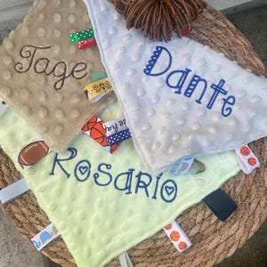 May include: Three square baby security blankets with dimpled textures in tan, white, and light green. Each blanket features embroidered names: Tage, Dante, and Rosario. The edges are adorned with various colourful fabric ribbon tags and loops.