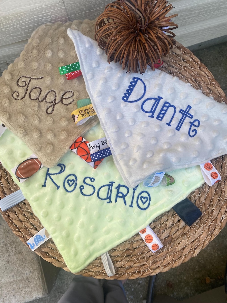 May include: Three square baby security blankets with dimpled textures in tan, white, and light green. Each blanket features embroidered names: Tage, Dante, and Rosario. The edges are adorned with various colourful fabric ribbon tags and loops.