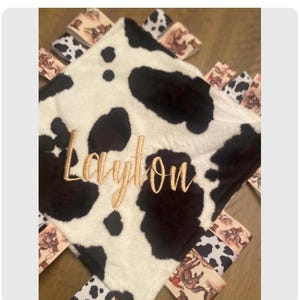 May include: A square security blanket featuring a black and white cow print pattern with gold embroidered text that reads Laylon. The edges are decorated with multiple fabric ribbon loops in cow print and western-themed cowboy illustrations.