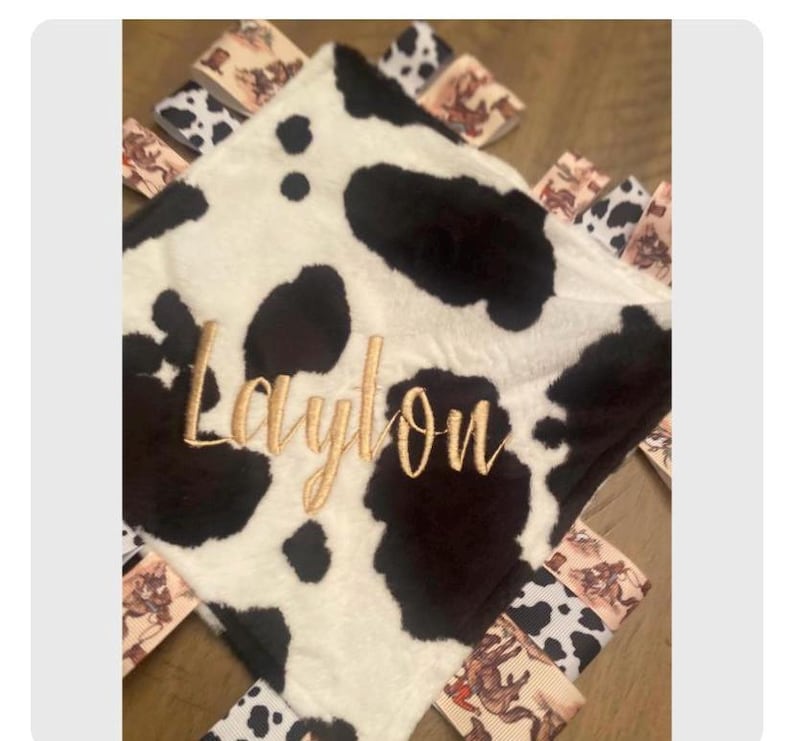 May include: A square security blanket featuring a black and white cow print pattern with gold embroidered text that reads Laylon. The edges are decorated with multiple fabric ribbon loops in cow print and western-themed cowboy illustrations.